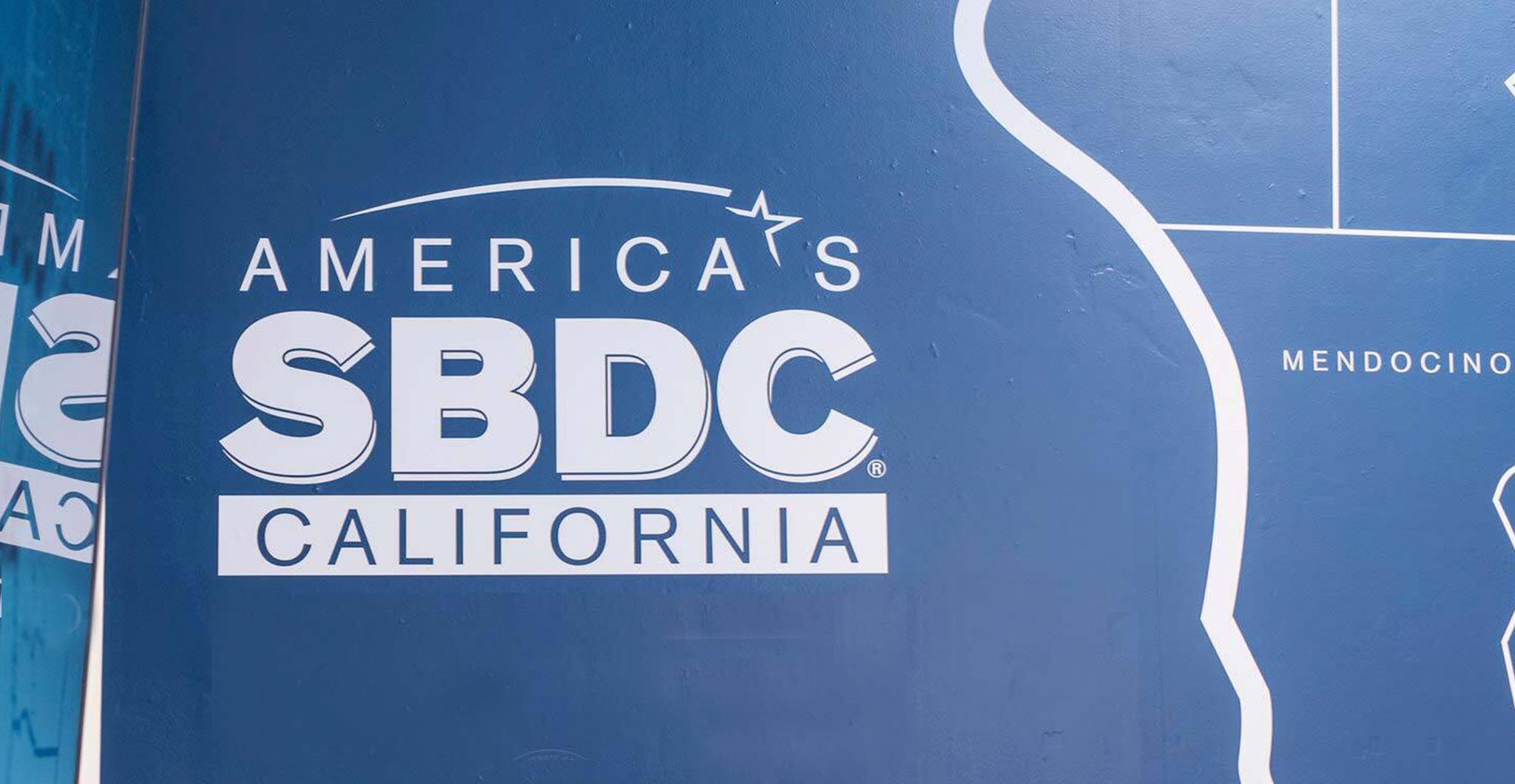 Close-up of the California SBDC identity and county map on a textured blue wall, with a reflection along the left edge.